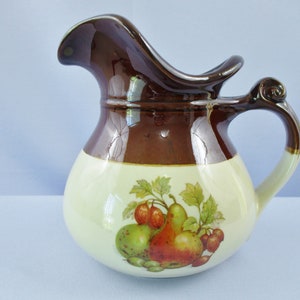 May include: A brown and cream ceramic pitcher with a wide spout and a handle. The pitcher has a fruit design with pears, apples, and berries.