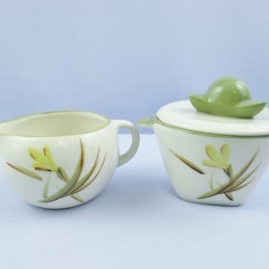 May include: A cream-colored ceramic creamer and sugar bowl set. The sugar bowl has a lid with a green handle. Both pieces are decorated with yellow floral designs and green and brown accents. The creamer has a handle and a spout.