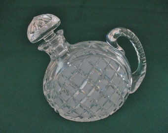 May include: A clear glass decanter with a diamond-shaped pattern and a round, crystal-like stopper. The decanter has a handle and is designed for serving beverages.