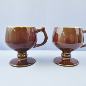 May include: Two brown ceramic mugs with a footed base and a handle. The mugs are glazed in a dark brown color.