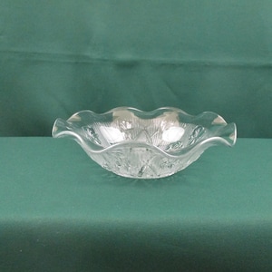 May include: A clear glass bowl with a scalloped rim and a decorative pattern. The bowl is empty and sitting on a green surface.