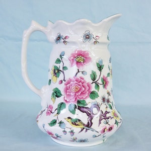May include: A white ceramic jug with a floral design featuring pink, yellow, and blue flowers and a small yellow bird. The jug has a scalloped rim and a handle.