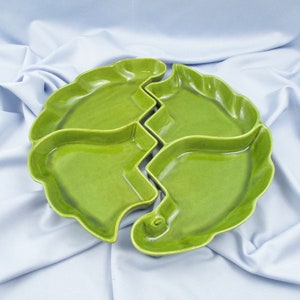 May include: A four-sectioned, leaf-shaped serving dish in a vibrant green hue. The dish is divided into four distinct sections, each with a gently curved edge. The sections fit together to form a circular shape. The dish is made of ceramic.
