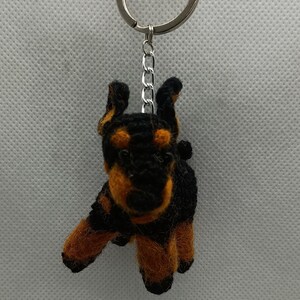 Puede incluir: Llavero hecho a mano con un pequeño perro Doberman Pinscher de peluche. El perro tiene un cuerpo negro con marcas marrones en las patas, el pecho y la cara. El llavero incluye un llavero y una cadena plateados para sujetar a llaves o bolsos.