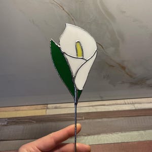 May include: A stained glass calla lily flower ornament featuring a white petal, a yellow spadix, and a dark green leaf, all held together by silver-colored metal solder lines on a thin metal stem.