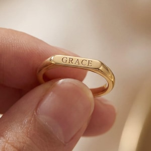 May include: A gold-colored ring with the word "GRACE" engraved on the top. The ring is held between fingers, showcasing its polished surface and simple design. The ring is a band style, with a rectangular top.