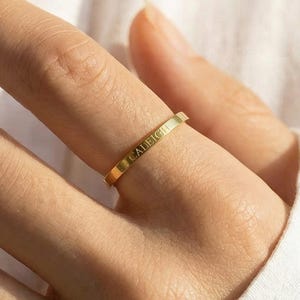 May include: A gold-colored ring with the engraved name "CALEIGH" is worn on a finger. The ring is thin and rectangular, with the text running horizontally. The background is a white shirt.
