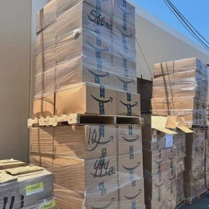 May include: Stacks of cardboard boxes, wrapped in clear plastic, are stacked on wooden pallets. The boxes have the Amazon logo and are labeled with handwritten numbers. The boxes are outdoors against a building and a blue sky.