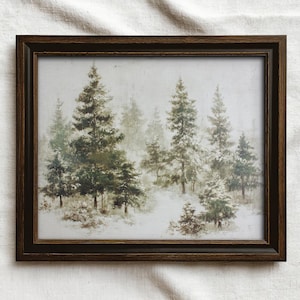 May include: A framed print featuring a winter forest scene. The artwork depicts evergreen trees covered in snow, with a muted color palette of greens, whites, and browns. The frame is a dark brown color.