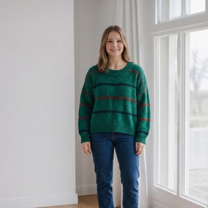 Könnte beinhalten: Ein langärmeliger Pullover mit Rundhalsausschnitt aus strukturiertem, smaragdgrünem Strick. Das Kleidungsstück hat horizontale Streifen in Dunkelmarineblau und Rostorange auf Brust und Ärmeln sowie gerippte Bündchen an Ärmeln und Saum, kombiniert mit einer blauen Jeans.