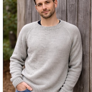 May include: A light gray sweater with a crew neck and raglan sleeves. The sweater is made of a knitted material and has a ribbed hem and cuffs. The person is leaning against a wooden fence.