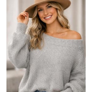Peut inclure: Un pull gris clair, épaules nues, à manches longues. Le pull a une texture en tricot côtelé et est associé à un chapeau fedora marron. La femme sourit et regarde la caméra.