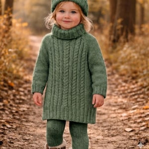 Op de afbeelding: Een kind in een groene gebreide outfit, bestaande uit een coltrui jurk, legging en bijpassende muts. De outfit heeft een kabelbreipatroon. Het kind staat op een bospad met herfstbladeren.