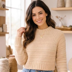 May include: A beige, cropped sweater with a ribbed crew neck and long sleeves. The sweater has horizontal textured stripes. A gold pendant necklace is visible. The person is smiling and wearing blue jeans.
