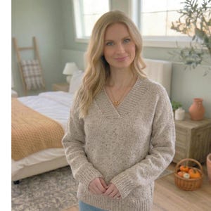 May include: A woman wearing a beige, speckled knit sweater with a V-neck. She is wearing a gold necklace with two interlocking circles. The background includes a bed with a yellow blanket, a wooden ladder, and a small table with a basket of pumpkins.