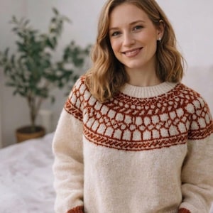 May include: A cream-coloured sweater featuring a geometric pattern in brown around the neckline and upper chest. The sweater has a crew neck and long sleeves with brown cuffs. The woman in the image is smiling.