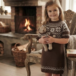 May include: A brown knit dress featuring a white snowflake and diamond pattern around the yoke and hem, paired with a matching knit teddy bear toy that wears a sweater with the same white snowflake and diamond pattern design.