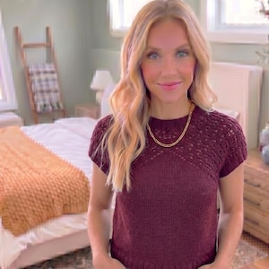 May include: A woman wearing a burgundy short-sleeved sweater with a gold chain necklace. The sweater has a textured, open-knit design on the upper chest and shoulders. The background includes a bed with a mustard-colored blanket and a wooden ladder.