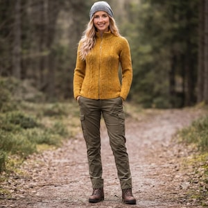 Op de afbeelding: Een mosterdgeel vest met kabelbreisel, ritssluiting en opstaande kraag, gecombineerd met een olijfgroene cargobroek met zijzakken, een grijze gebreide muts en bruine leren wandelschoenen op een bospad.