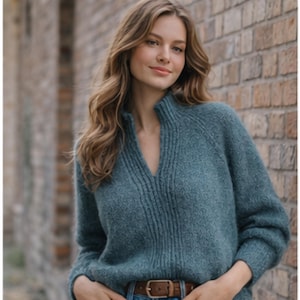 May include: A teal-colored sweater with a V-neckline and ribbed detailing. The sweater has long sleeves and a relaxed fit. The model is wearing a brown belt with a gold buckle. The background is a brick wall.