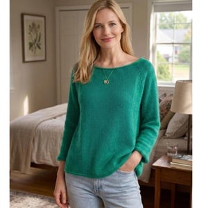 Può includere: Una donna indossa un maglione verde acqua con scollo a barchetta e maniche lunghe. Indossa una collana d'oro e jeans azzurri. Il maglione è di colore pieno e sembra essere realizzato in un materiale morbido e lavorato a maglia. Lo sfondo include un letto, una lampada e una finestra.