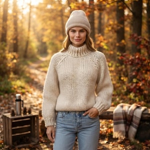 Pode incluir: Uma camisola de gola alta em malha grossa de cor creme com mangas compridas e um gorro de malha canelada a combinar, usada com calças de ganga azul-claras. A camisola apresenta uma trama espessa e texturizada com uma gola alta.