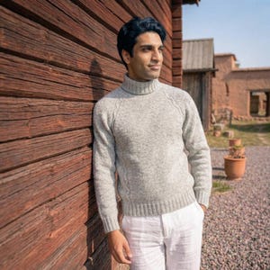 Puede incluir: Un suéter de cuello alto gris claro de manga larga con un sutil patrón de trenzas en las mangas y los costados, combinado con pantalones blancos. El suéter tiene cuello, puños y dobladillo acanalados, confeccionado en tejido de punto jaspeado.