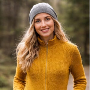 May include: A woman wearing a gray knit beanie and a mustard yellow zip-up sweater. The sweater has a high collar and a gold zipper. The woman has long blonde hair and is smiling. The background is blurred, with green and brown tones.