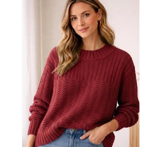 May include: A burgundy-colored sweater with a ribbed crew neck and cuffs. The sweater features a textured, patterned design on the front and sleeves. The model is wearing blue jeans.