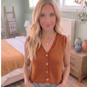 May include: A sleeveless, rust-colored knit vest featuring a V-neckline, a front button-down closure with four light-colored buttons, and a scalloped hemline. The garment is constructed from a textured knit fabric with a ribbed trim along the neckline.