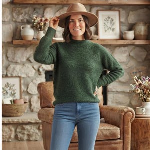 Puede incluir: Un suéter de manga larga color verde oscuro con un patrón de tejido texturizado y cuello alto, combinado con jeans azul claro y un sombrero de fieltro color canela de ala ancha. El suéter presenta un corte ajustado con puños y dobladillo acanalados.