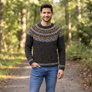 Pode incluir: Uma camisola de malha de manga comprida em castanho escuro com decote redondo e um padrão decorativo no jugo em tons de azul, laranja e castanho. A camisola é combinada com calças de ganga azuis, com um acabamento de textura semelhante à lã.