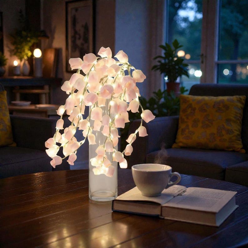 May include: A clear glass vase holds a cascading arrangement of illuminated pink flower lights. A cup of coffee sits on a wooden table next to an open book. The scene is set in a cozy living room with soft lighting and a dark gray sofa.