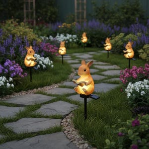 Könnte beinhalten: Solarbetriebene Gartenleuchten in Form von Hasen, die Bücher lesen. Die Leuchten sind orange und gelb und strahlen ein warmes Licht aus. Sie sind entlang eines Steinwegs in einem Garten mit bunten Blumen platziert.