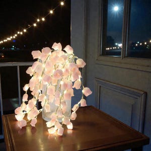May include: A glass vase holds a cascade of soft pink, bell-shaped fairy lights, creating a warm glow. The lights are arranged on a dark wooden table, with a window and string lights in the background.