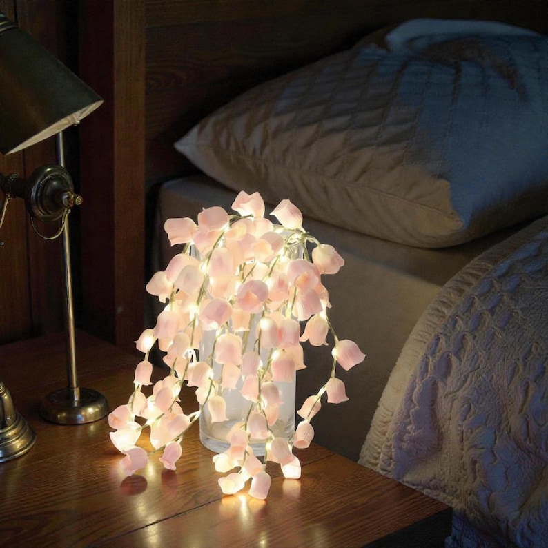 May include: A glass vase filled with cascading pink flower string lights. The lights are on, illuminating the delicate petals. The vase sits on a wooden nightstand next to a bed with a white pillow and comforter.