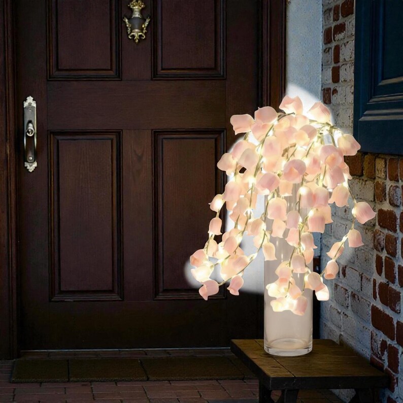 May include: A decorative floral light string with soft pink flower-shaped lights cascading from a clear glass vase. The lights are on a dark wooden table, next to a brick wall and a dark brown door.