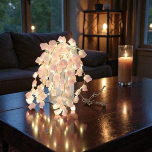 May include: A string of fairy lights shaped like pink flowers cascading from a clear vase on a dark wooden table. A lit candle in a glass cylinder sits nearby. The scene is softly lit, creating a warm ambiance.