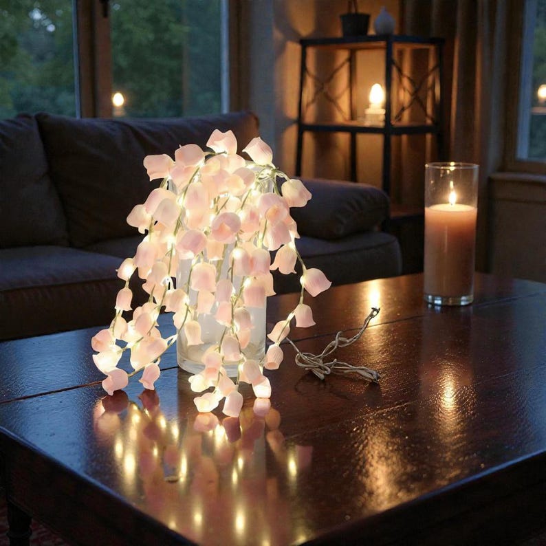 May include: A string of fairy lights shaped like pink flowers cascading from a clear vase on a dark wooden table. A lit candle in a glass cylinder sits nearby. The scene is softly lit, creating a warm ambiance.