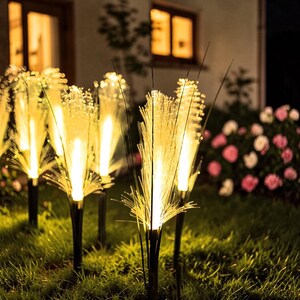 Op de afbeelding: Zonne-energie tuinverlichting met glasvezel. De lichten staan op een gazon, elk met een zwarte basis en een cluster van verlichte glasvezelstrengen, die een warme, gouden gloed uitstralen. Op de achtergrond is een bloembed te zien.