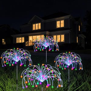 Op de afbeelding: Vier decoratieve buitenlampen met witte stelen en veelkleurige LED-verlichting. De lichten zijn gerangschikt in een cluster, die op vuurwerk lijken. De lichten staan 's nachts in een grasveld voor een huis.