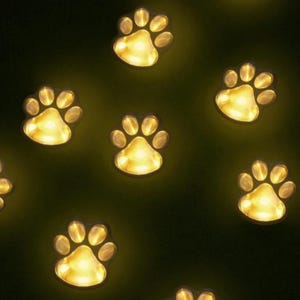 Puede incluir: Un patrón de luces brillantes en forma de huellas de patas sobre un fondo oscuro. Cada huella emite una luz cálida y dorada, creando un ambiente caprichoso y acogedor. Las luces están espaciadas uniformemente, lo que sugiere una aplicación decorativa o festiva.