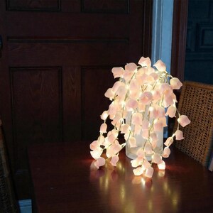 May include: A string of fairy lights with soft pink flower-shaped shades cascading from a clear glass vase. The lights are illuminated, creating a warm, inviting glow. The arrangement sits on a dark wooden surface.