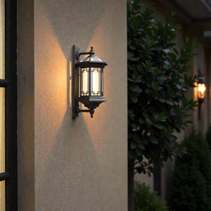 Puede incluir: Una linterna negra de exterior, montada en la pared, con un diseño rectangular y una parte superior decorativa. La linterna tiene un panel de vidrio que emite una luz cálida, proyectando una sombra en la pared beige. Una segunda linterna es visible en el fondo.