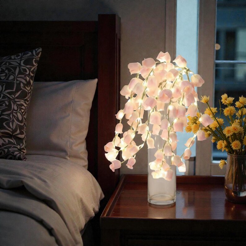 May include: A decorative arrangement of pink flower petals with small lights in a clear glass vase. The vase sits on a dark brown nightstand next to a bed with a patterned pillow and a vase of yellow flowers.