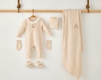 May include: A beige baby clothing set, including a onesie, hat, blanket, mittens, and booties. The onesie has a white lace collar and floral embroidery. The blanket features a matching embroidered design. The set is displayed on a wooden rod.