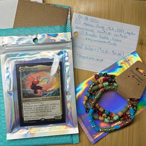 May include: An Avatar Aang trading card in a clear plastic sleeve, alongside a bracelet bundle with colorful beads. The card features a character illustration. The items are on a wooden surface.