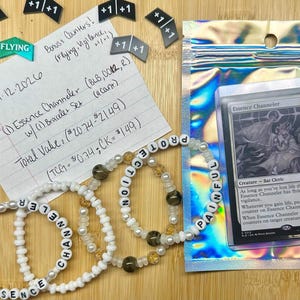 Puede incluir: Un surtido de artículos que incluyen pulseras de cuentas que deletrean "ESSENCE CHANNELER" y "PROTECTION PAINFUL", contadores de juego y una carta en una funda protectora. La carta está etiquetada como "Essence Channeler". También hay una nota manuscrita.