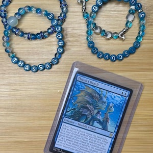 May include: Two beaded bracelets featuring blue and clear glass beads with silver-toned accents and letter beads spelling THASSA'S ORACLE and DEVOTION DISCOVERY, alongside a Thassa's Oracle trading card encased in a clear protective plastic sleeve.