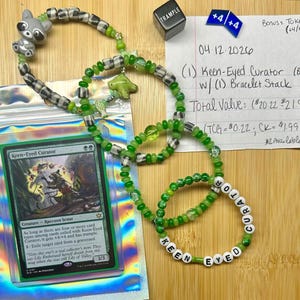 Puede incluir: Una pulsera de cuentas verdes y blancas con el texto "KEEN EYED CURATOR" se muestra con una carta coleccionable y otros accesorios. La tarjeta se titula "Keen-Eyed Curator". También se ve una pequeña cuenta de mapache gris.